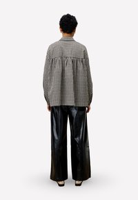 Persoon met kort krullend haar, gekleed in een oversized zwart-wit geblokt shirt en wijdlopende zwarte leren broek, staand met de rug naar de kijker.