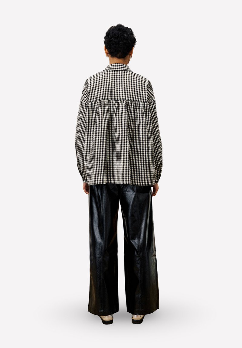 Persoon met kort krullend haar, gekleed in een oversized zwart-wit geblokt shirt en wijdlopende zwarte leren broek, staand met de rug naar de kijker.