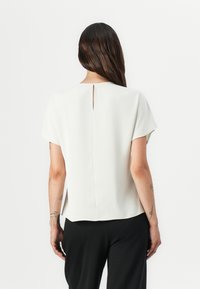 Witte blouse met korte mouwen, voorzien van een kleine opening aan de achterkant en gemaakt van een soepele, lichte stof. Gepaard met zwarte broeken.