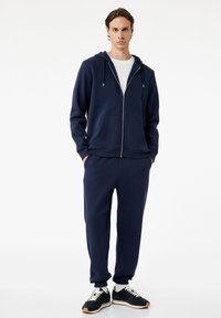 Sudadera con cremallera de color azul marino con cordones, combinada con pantalones jogger a juego. Cuenta con un bolsillo frontal y puños elásticos. Zapatillas negras con acentos blancos.