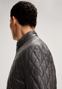 Mann, der sich abwendet und eine dunkelbraune, gesteppte Lederjacke mit runder gestickter Applikation am oberen Ärmel trägt.