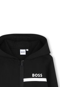 Sweat à capuche noir zippé avec capuche, arborant un logo "BOSS" blanc au-dessus d'une bande blanche horizontale sur la poitrine.