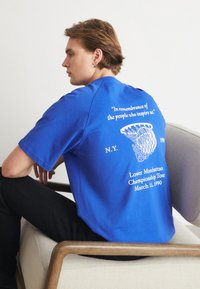 Zila kokvilnas T-kreklis ar īsām piedurknēm, aizmugurē ar baltu tekstu un basketbola groza grafiku. Teksts ietver notikuma detaļas.