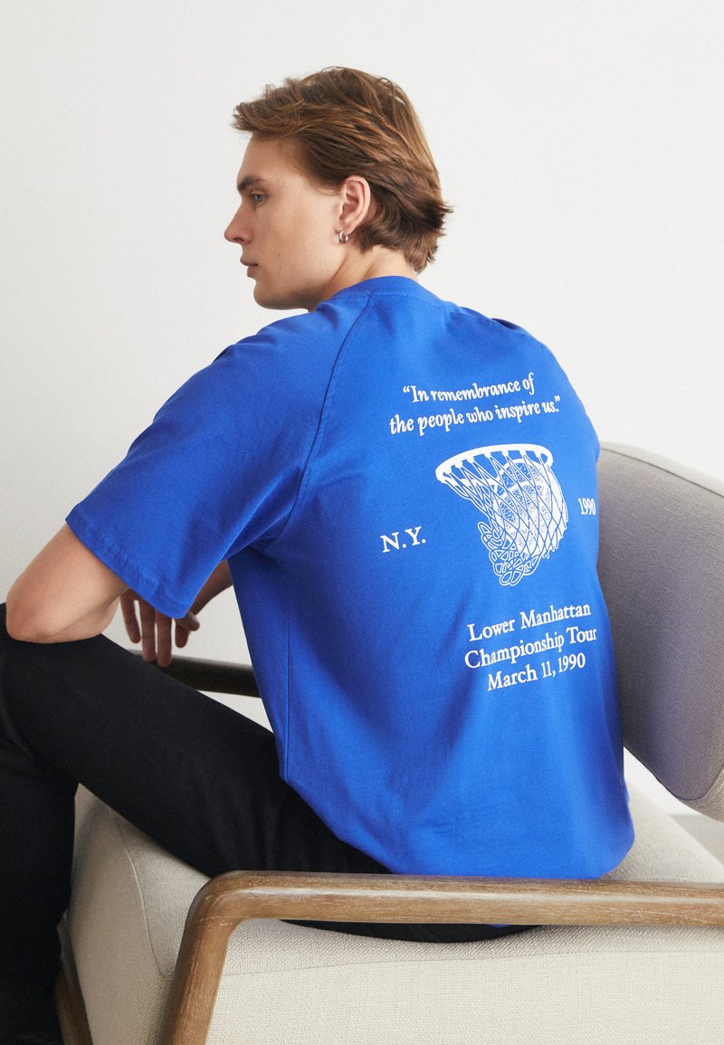 Modré bavlněné tričko s krátkým rukávem, na zadní straně s bílým textem a grafikou basketbalového koše. Text obsahuje podrobnosti o akci.