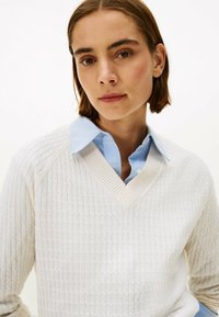 Pull en V crème avec un motif de tricot texturé, superposé sur une chemise col bleu clair. Présente des poignets et un col côtelés.