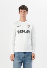 Ung mand iført en hvid langærmet skjorte med "REPLAY" og et våbenskjoldlogo, kombineret med blå jeans, stående foran en hvid baggrund.