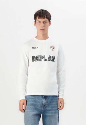Young man wearing a white long-sleeve shirt with "REPLAY" and a crest logo, paired with blue jeans, standing against a white background.