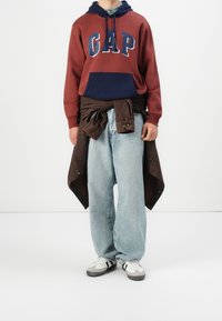 Tummanpunainen ja laivastonsininen GAP-huppari, väljät vaaleansiniset farkut, ruskea takki sidottuna vyötärölle ja valkoiset urheilukengät harmailla yksityiskohdilla.