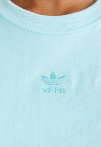 Ljusblå bomullströja med ribbad halsringning och subtilt broderad Adidas-logotyp i mitten, med en tre-randers design ovanför texten.