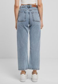 Jeans de tiro alto de lavado claro en azul, con corte de pierna amplia, dos bolsillos traseros y dobladillo sin rematar. Combinados con zapatillas blancas. Textura de denim suave.