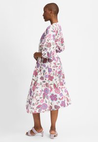 Ein florales Maxikleid in Weiß mit pinken und blauen Mustern, ausgestattet mit Puffärmeln, einer taillierten Taille und einer fließenden Silhouette. Kombiniert mit lila High Heels.