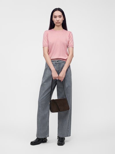 Jeune femme aux longs cheveux noirs portant un t-shirt rose à manches courtes, un jean gris à jambes larges, des bottes noires et tenant un sac à main marron.