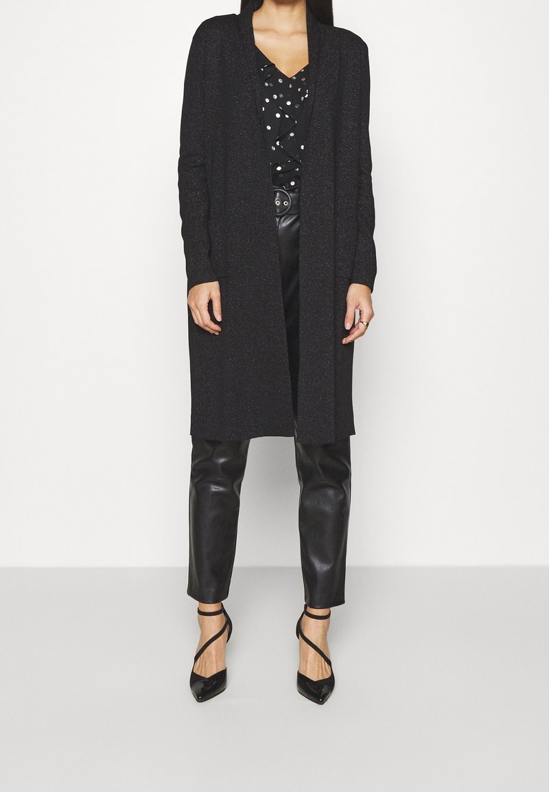 Black long cardigan with open front, ribbed texture, paired with a black and white polka dot blouse, black faux leather pants, and black heels.