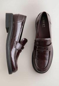 Mocassini in pelle laccata color bordeaux con finitura lucida, design slip-on e suola in gomma nera. Presentano una fascia decorativa nella parte anteriore.