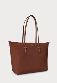 Καφέ τσάντα ώμου με υφή, με δύο μακριές δερμάτινες λαβές, λογότυπο σε χρυσό τόνο και κλείσιμο με φερμουάρ στην κορυφή, σε απλό ανοιχτόχρωμο φόντο.