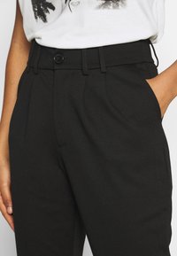 Pantalon noir à la coupe ajustée, avec fermeture à bouton, poches latérales et plis visibles. Le tissu semble lisse et structuré.