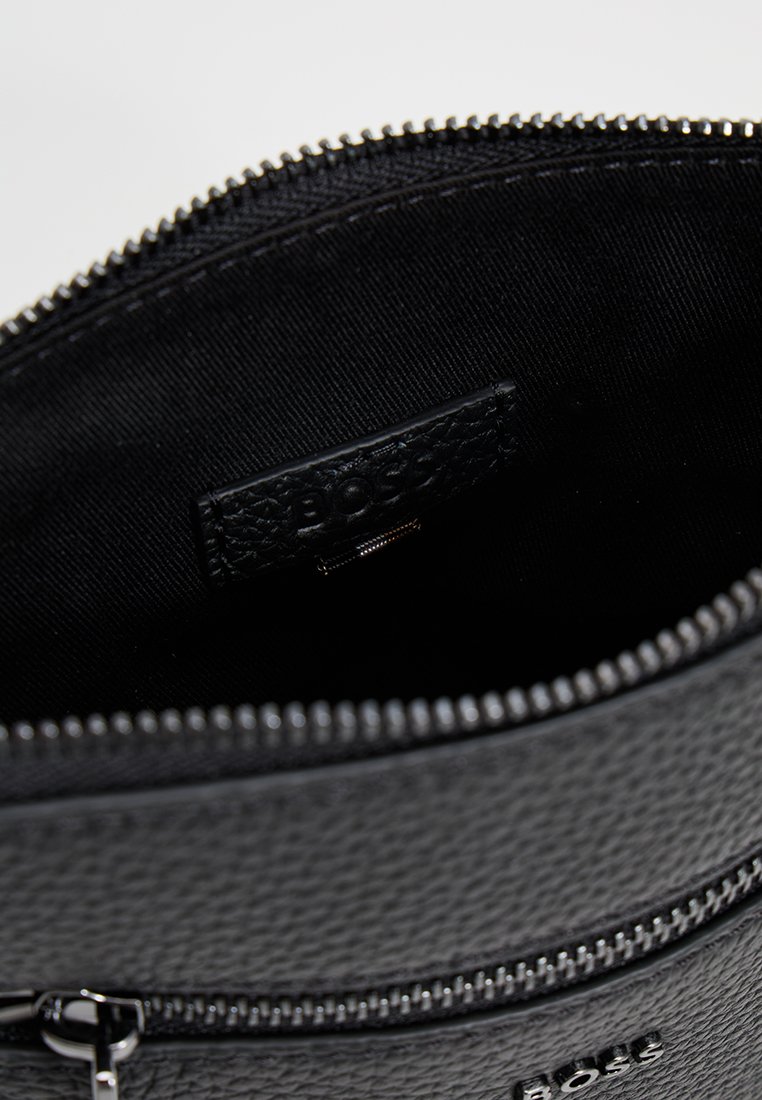 Pochette de voyage en cuir noir avec une surface texturée, dotée d'une fermeture éclair, et d'un intérieur en tissu noir avec une étiquette logo.