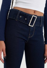 Mørkeblå denimjeans med et bredt belte som har en rektangulær sølvspenne, syddetaljer og en tettsittende passform rundt midjen og lårene.