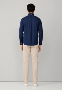 Chemise bleu marine à manches longues avec un col boutonné, vue de l'arrière ; associée à un pantalon beige clair et des baskets blanches.