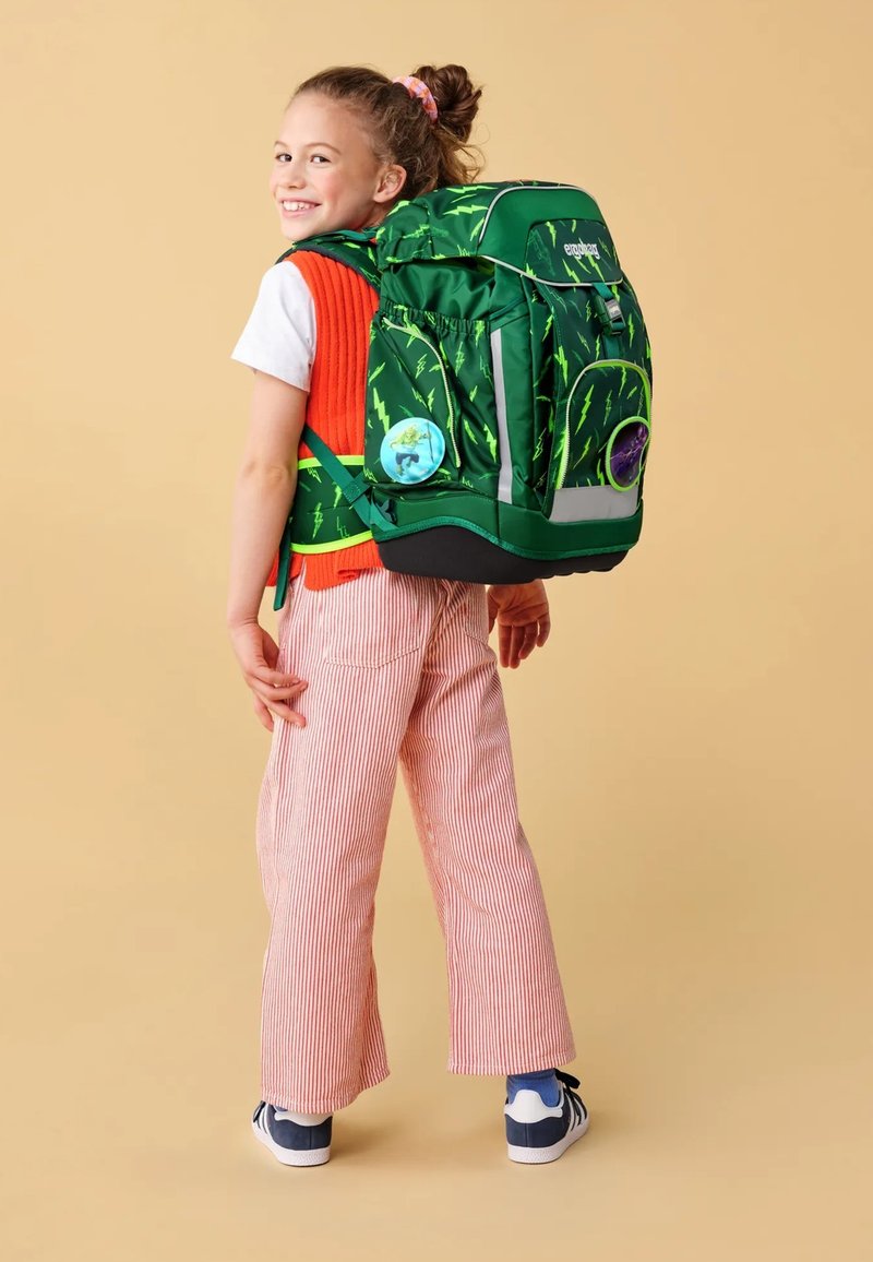 Grüner Rucksack mit Blitzdesign, reflektierenden Akzenten, Seitentaschen und runden Patches; getragen über einem orangenen Pullover und gestreiften Hosen.