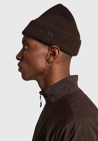 Brązowa prążkowana dzianinowa czapka z haftowanym logo na opasce, prezentowana na osobie noszącej kurtkę z wysokim kołnierzem i kolczykiem.