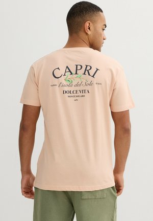 Hombre vistiendo una camiseta color melocotón con el texto "CAPRI l'isola del Sole Dolce Vita" y pantalones cortos verde oliva, de espaldas sobre un fondo liso y claro.