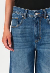 Blue denim high-waist jeans feature a front button closure, contrast stitching, and side pockets. Texture appears smooth with slight fading.
