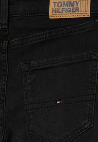 Pantalones vaqueros negros con una etiqueta beige que dice "TOMMY HILFIGER". Pequeño logo bordado en rojo, blanco y azul en el bolsillo trasero. Textura suave.