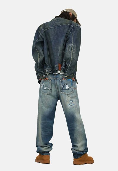 Persona che indossa una giacca di jeans sbiadita e jeans larghi con disegni cuciti sulle tasche, stivali marrone chiaro e un berretto da baseball beige, in piedi con le mani nelle tasche.