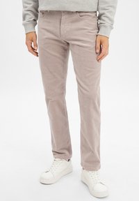 Man in lichtbeige corduroy broek en witte sneakers, staand tegen een plain witte achtergrond.