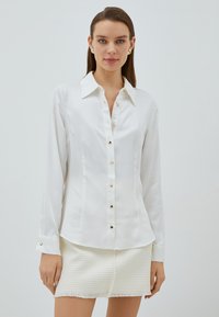 Blusa bianca a maniche lunghe con colletto a punta, caratterizzata da dettagli con bottoni dorati e una texture liscia. Abbinata a una gonna bianca con texture.
