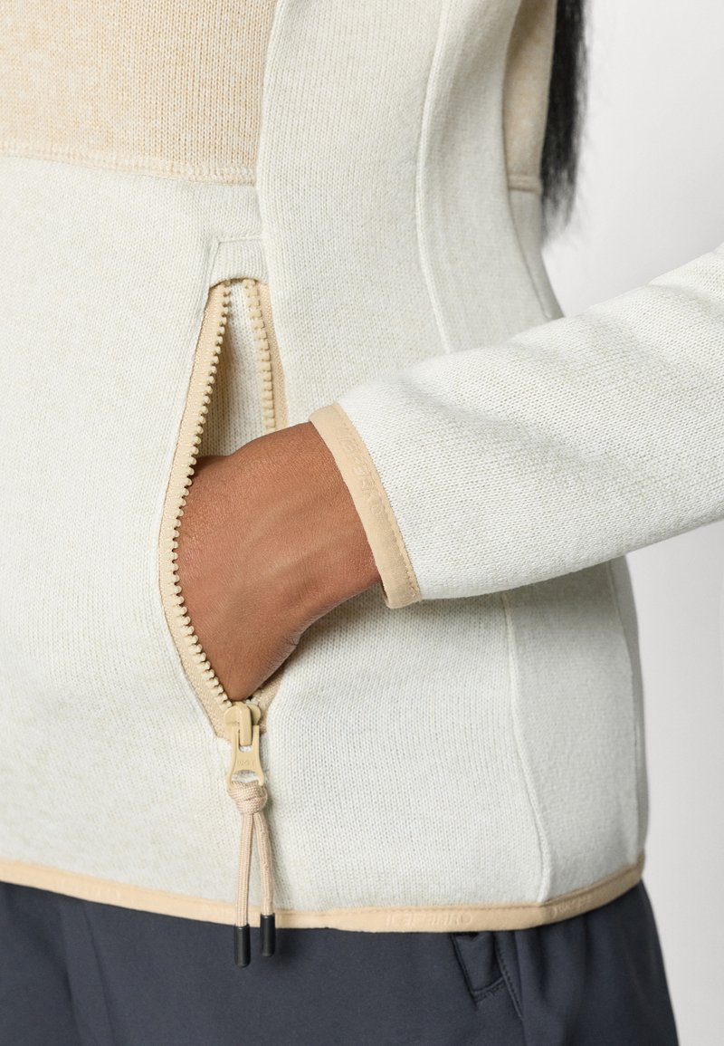 Sweater con cremallera de color claro, con tela texturizada y acentos en beige, y una mano reposando en el bolsillo lateral. El tirador de la cremallera es beige con puntas negras.