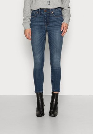 Kobieta w niebieskich dopasowanych dżinsach, szarej bluzie z literą B i czarnych botkach, stojąca na jednolitym jasnym tle.