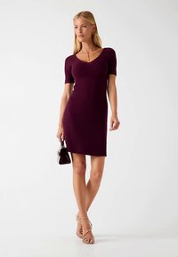 Donkerpaarse, geribbelde jurk met V-hals, korte mouwen en aansluitende pasvorm; model houdt een kleine zwarte handtas vast en draagt sandalen met bandjes in een nude kleur.