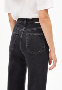 Jean en denim noir à taille haute, avec des poches arrière et une étiquette de marque visible. Tissu texturé avec des détails de décoloration subtils.