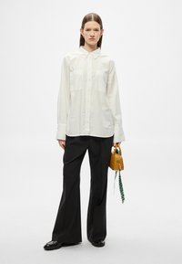 Fehér, gombos ing két mellzsebbel, fekete, széles szárú nadrággal párosítva. A modell egy kis sárga táskát tart zöld pánttal.