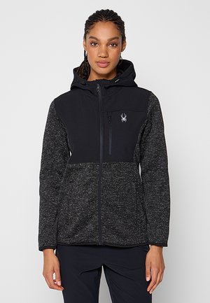 Femme portant un sweat à capuche zippé noir et gris foncé avec un logo d'araignée sur la poitrine, se tenant contre un fond clair uni.
