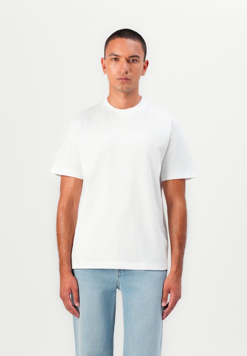 Weißes Baumwoll-T-Shirt mit regular Fit, Rundhalsausschnitt, kurzen Ärmeln und einem dezenten strukturierten Logo auf der Vorderseite. Kombiniert mit hellen Jeans.