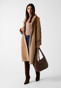Beżowy długi płaszcz z owczej wełny z kołnierzem, z prążkowanymi mankietami, w połączeniu z jasnym golfem, ciemnymi dżinsami, brązowymi zamszowymi kozakami do kolan oraz brązową torebką.