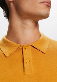 Senfgelbes Poloshirt mit strukturiertem Strickdesign, Kragen und zwei Knöpfen. Glatte Stofffläche am Halsauschnitt sichtbar.