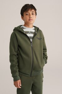 Zelená mikina na zips vyrobená z mäkkého materiálu, s logom na hrudi, klokaničími vreckami a rebrovanými manžetami, nosená cez pruhované tričko.