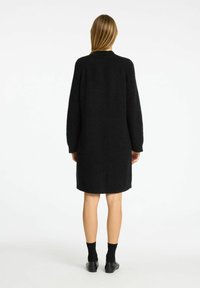 Schwarzer gestrickter Mantel mit lockerer Passform, langen Ärmeln und mittellanger Silhouette. Weicher Stoff mit geripptem Kragen und ohne sichtbare Verschlüsse.