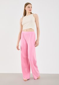 Lyserøde bukser med bred benvidde og glat tekstur, parret med en tæt siddende cremegul ribbet crop-top. Modellen står foran en neutral baggrund.