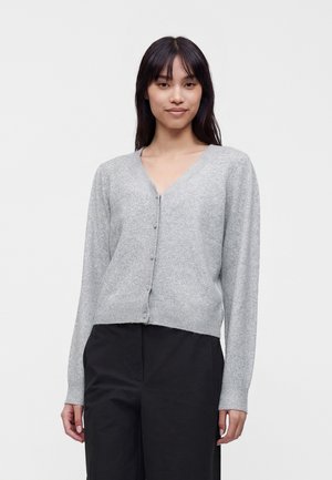 Femme portant un cardigan gris clair boutonné et un pantalon noir, debout devant un fond blanc uni.