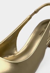 Χρυσό σατέν παπούτσι με λουράκι πίσω, στρογγυλεμένη μύτη και μινιμαλιστικό σχέδιο. Διαθέτει ομαλή υφή και διακριτικό τακούνι για επιπλέον ανύψωση.