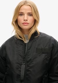 Sort bomberjakke med lynlås foran, ribbet krave og patch-detaljer. Har en tekstureret stof og diskrete logo-accenter.
