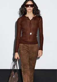 Braunes, geripptes Langarmoberteil kombiniert mit braunen Wildlederhosen, ergänzt durch dunkle Sonnenbrillen und eine braune Handtasche. Glatte Texturen, figurbetontes Design.