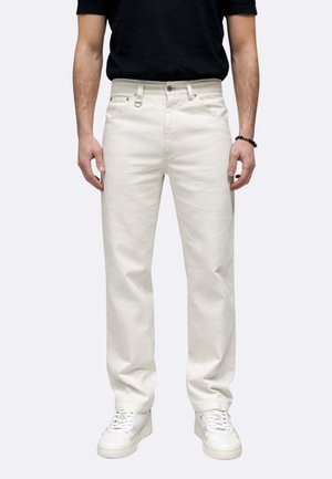 Uomo che indossa jeans dritti color avorio, sneakers bianche, braccialetto nero con perline e camicia nera a maniche corte, in piedi contro uno sfondo semplice.