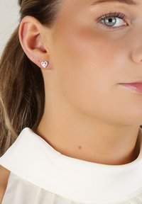 Silver hjärtformade örhängen med en polerad yta och små zirkon-accenter, bärs i ett hållet öra mot en ljusfärgad tyg.