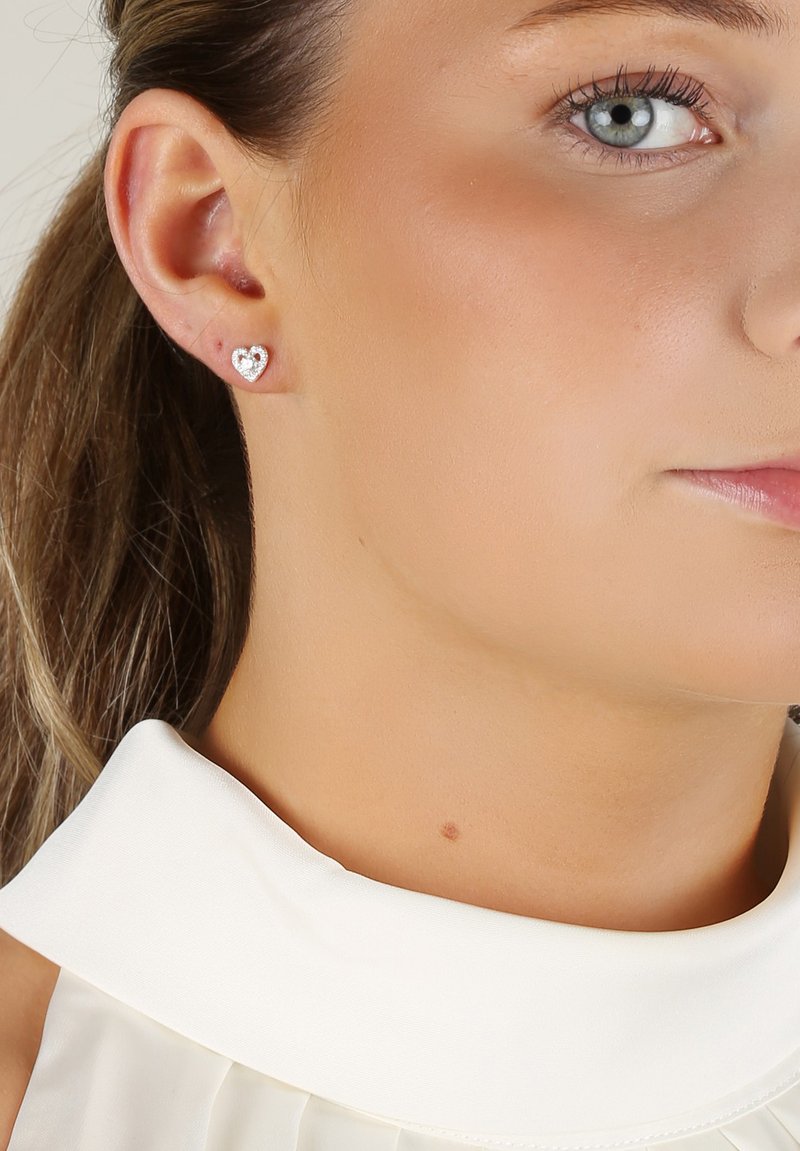 Silver hjärtformade örhängen med en polerad yta och små zirkon-accenter, bärs i ett hållet öra mot en ljusfärgad tyg.